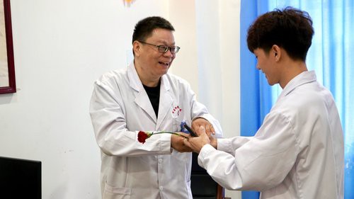 诊室桌旁，一位戴眼镜的医生笑着接过康乃馨，与一位年轻医生双手传递，背景是浅蓝色窗帘